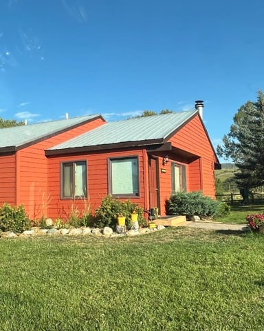 Red cottage with metal roof surrounded by evergreen trees and green lawn under clear blue sky