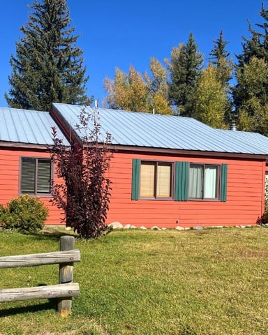Red cabin with metal roof surrounded by evergreen and aspen trees, wooden fence in foreground