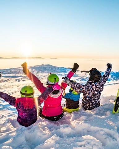 Four skiers in colorful winter gear celebrating on snowy mountain slope at sunrise with distant peaks visible