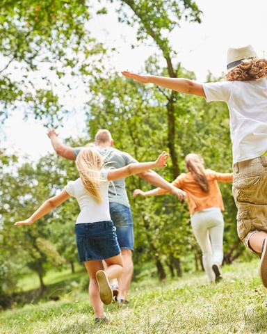 Family of four running through a grassy field surrounded by tall green trees on a sunny day.