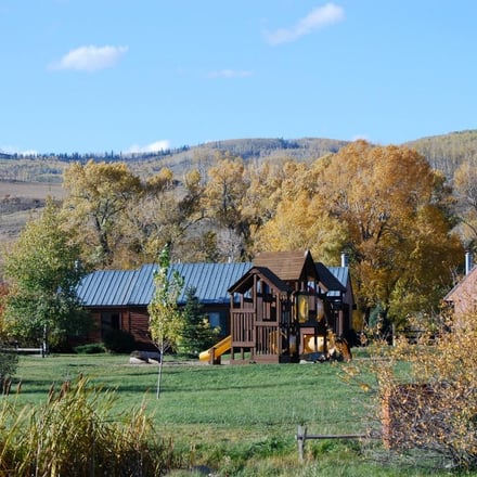 Log cabins with metal roofs and playground in grassy valley surrounded by autumn trees and forested hills