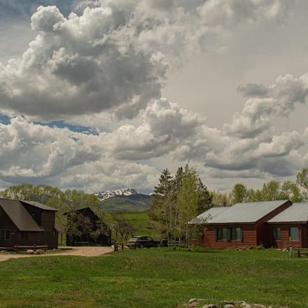 Two red-brown cabins with metal roofs on a grassy property, mountains visible in the distance under dramatic white clouds