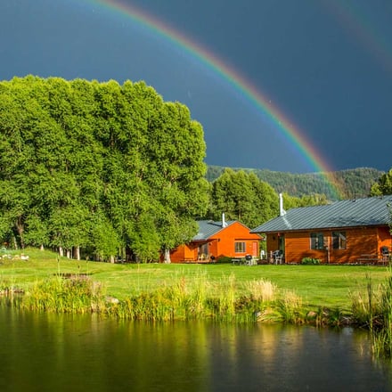 Bright orange log cabins on grassy property beside pond with large trees and rainbow in stormy sky