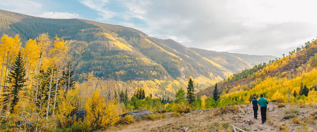 Two hikers on a rocky trail surrounded by golden aspen trees and evergreens in a mountain valley