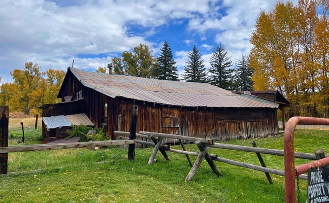 Weathered dark wooden cabin with metal roof surrounded by autumn trees and rustic wooden fence