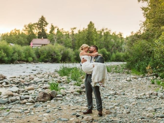 Groom carrying bride on rocky riverbank with forested mountains and cabin in background