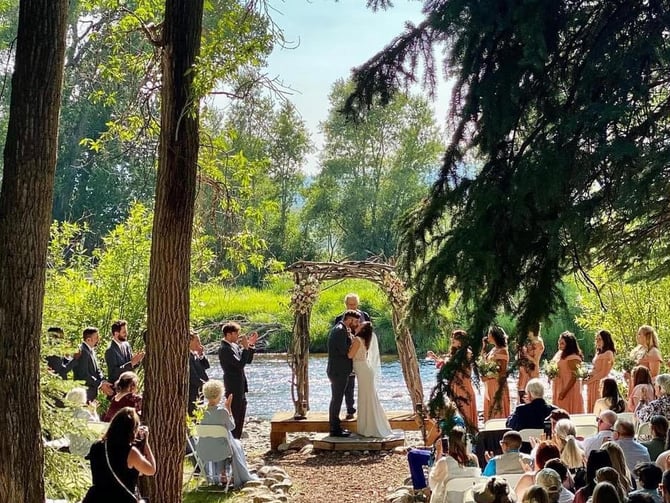 Outdoor wedding ceremony under tall trees beside a river, with bride and groom at wooden arch and guests seated on both sides.