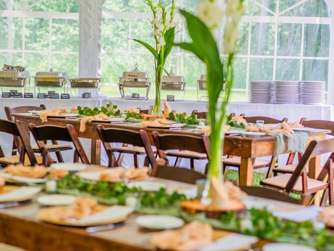 Banquet hall with wooden chairs, long tables set with food and flowers, warming trays, and large windows overlooking green landscape.