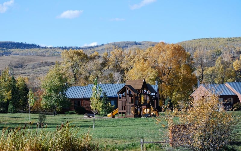 Log cabins with metal roofs and playground in grassy valley surrounded by autumn trees and forested hills