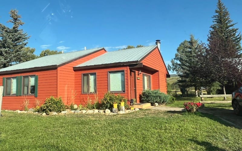 Red cottage with metal roof surrounded by evergreen trees and green lawn under clear blue sky