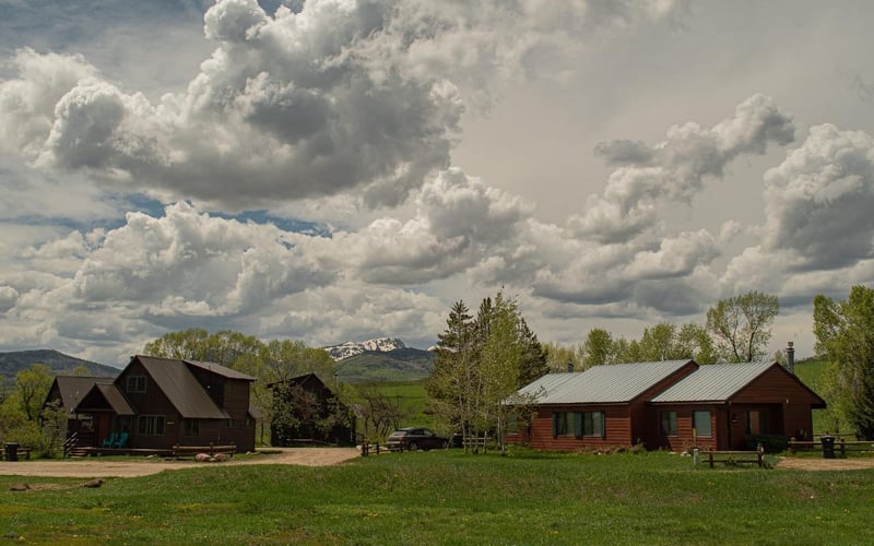 Two red-brown cabins with metal roofs on a grassy property, mountains and dramatic clouds in background.
